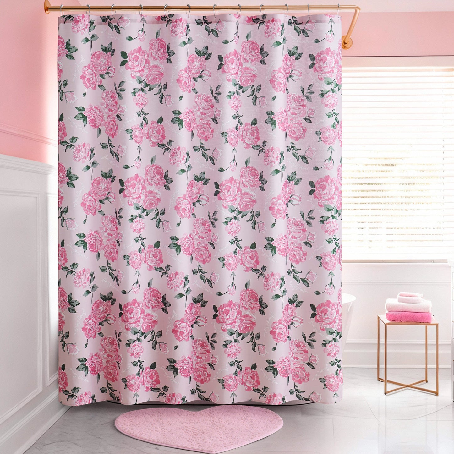 A pink and white bathroom features a Shower Curtain and Hooks Set with a pink rose floral pattern, a pink heart-shaped bath mat, and a stack of pink towels on a white stool by the window—stylish decor for any space.