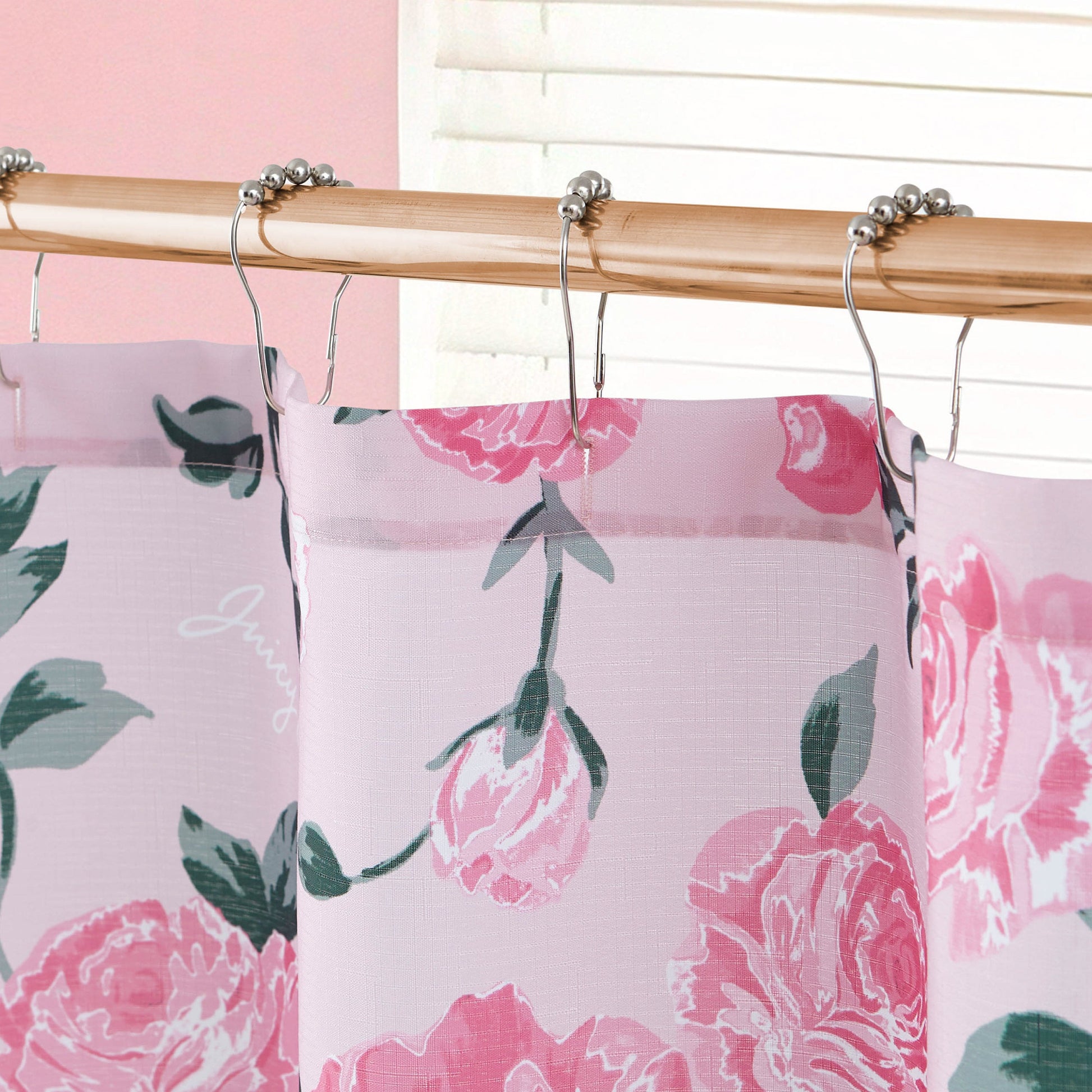 A close-up of the Shower Curtain and Hooks Set featuring pink florals and green leaves hangs on a bamboo rod with metal hooks, complemented by blinds and a light pink wall for a charming bathroom backdrop.