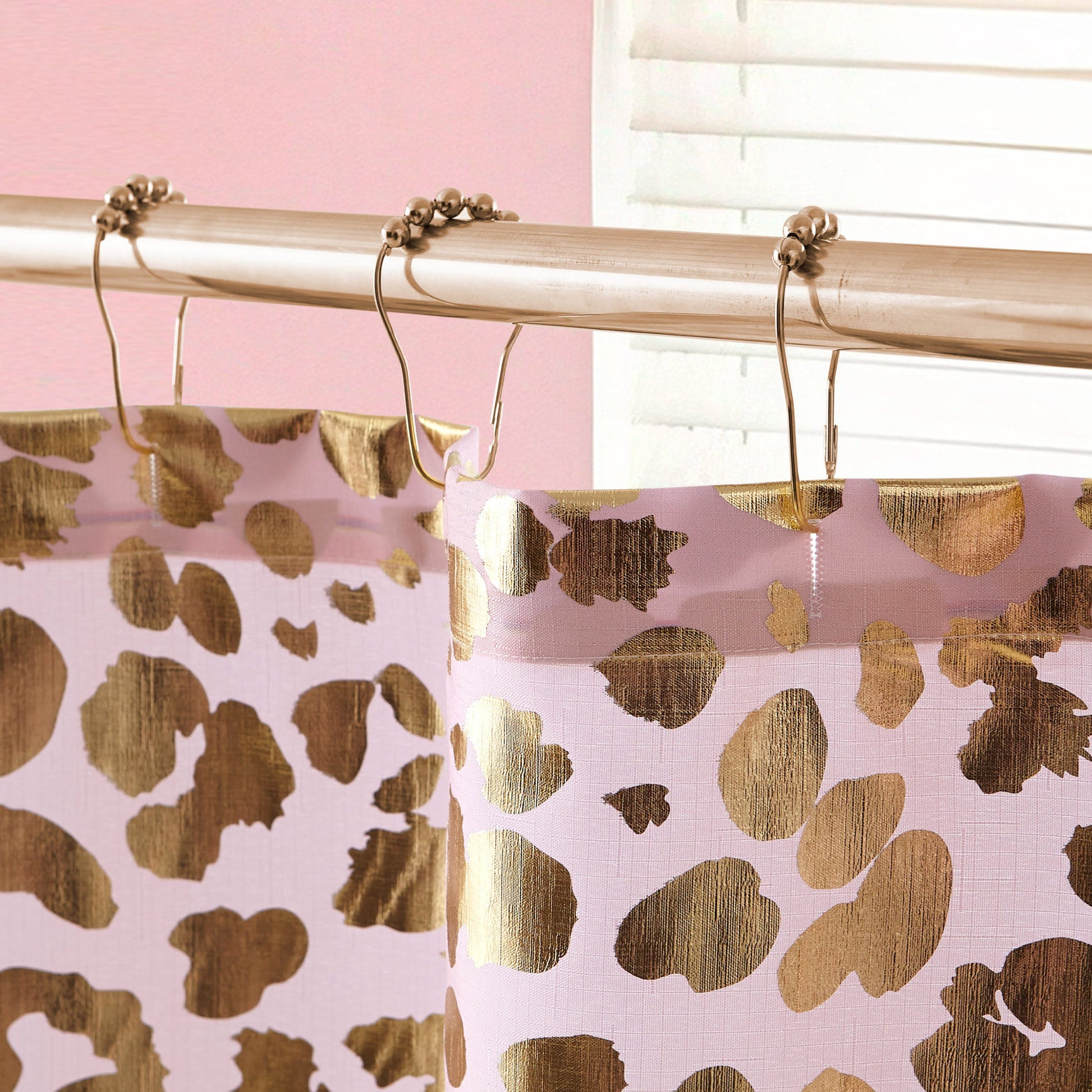 Close-up of the Shower Curtain and Hooks Set featuring a gold and pink leopard print, hanging from a silver rod with metal rings. The background shows stylish bathroom decor, including a pink wall and partially open white blind.