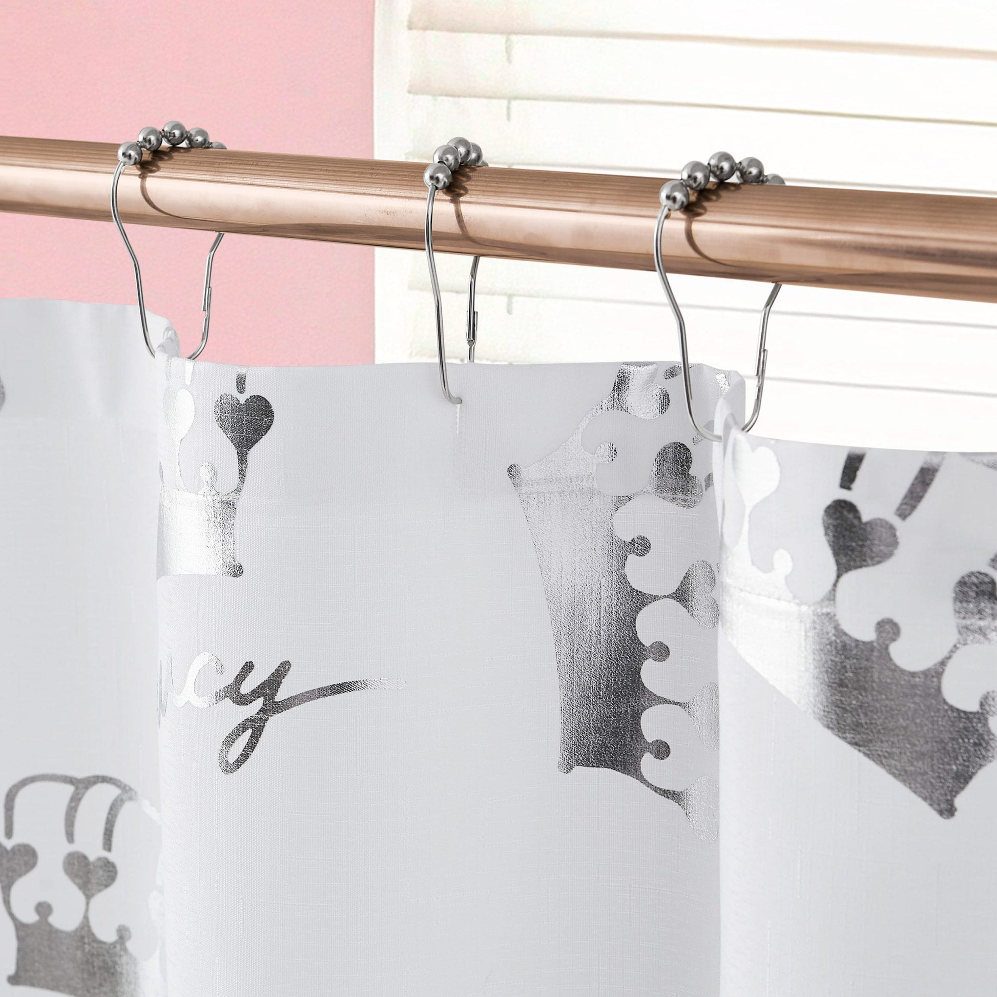 A close-up of the Shower Curtain and Hooks Set, featuring metallic silver crown designs. Hung on a rod with matching hooks, it’s set against a pink wall and white blinds for a chic bathroom accent.