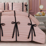 A neatly made bed with the Amelie Bows Ruched Comforter Set—pale pink bedding with black ribbon and velvet bows—sits by a mirrored nightstand holding a round mirror, perfume, and small items in a pink-walled room.