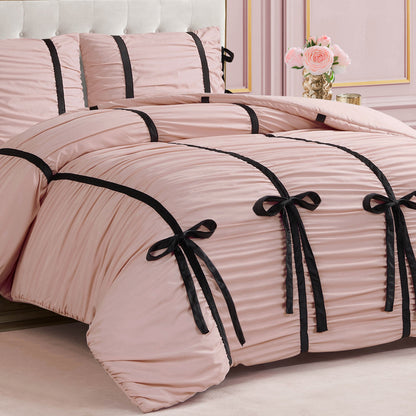 A neatly made bed with the Amelie Bows Ruched Comforter Set in pink, featuring velvet bows and black ribbon accents. A pink wall and a vase of pink roses on a table appear in the background.