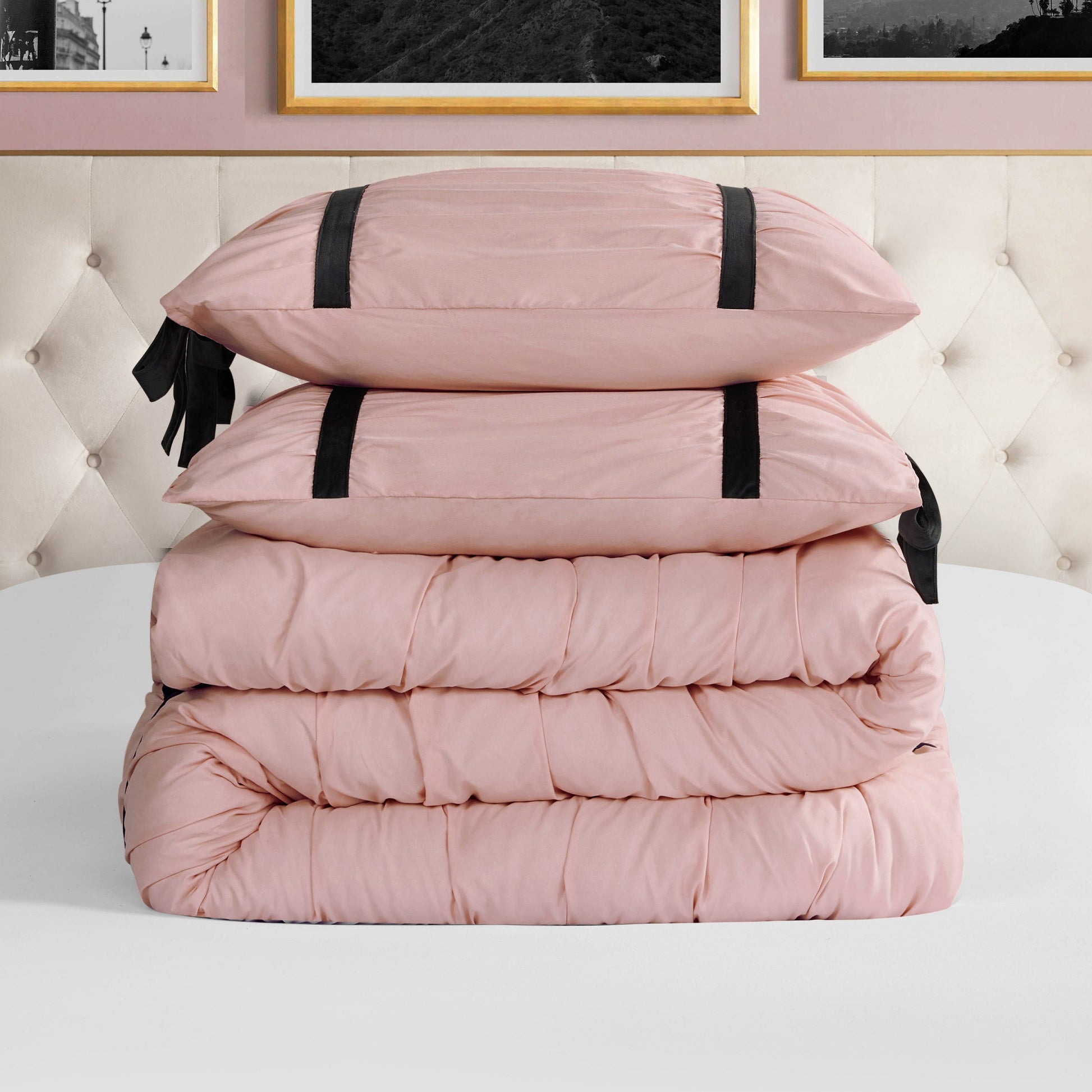 A neatly folded light pink Amelie Bows Ruched Comforter Set with two matching pillows featuring black velvet bows sits on a white surface before a tufted beige headboard and framed wall art.