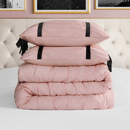 A neatly folded light pink Amelie Bows Ruched Comforter Set with two matching pillows featuring black velvet bows sits on a white surface before a tufted beige headboard and framed wall art.