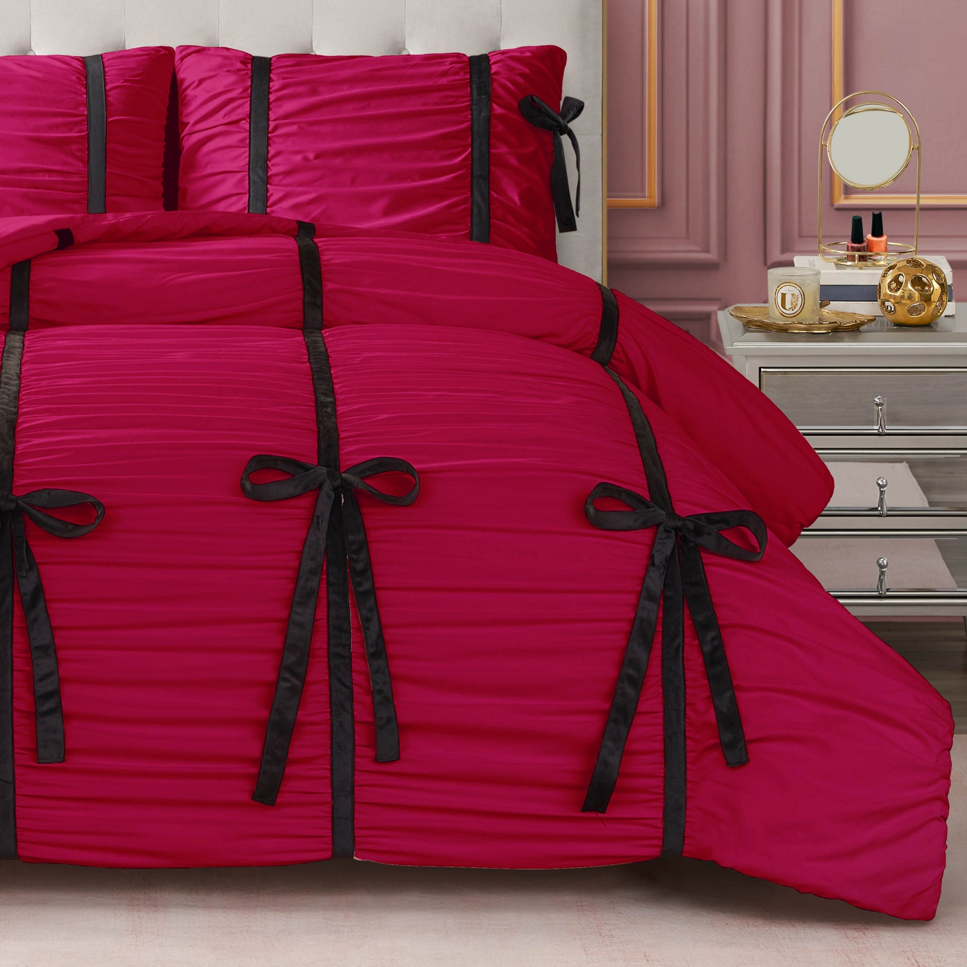 A bed with the Amelie Bows Ruched Comforter Set in bright pink, featuring vertical black velvet bows, stands next to a mirrored nightstand topped with a round mirror, cosmetics, and a gold decorative piece.