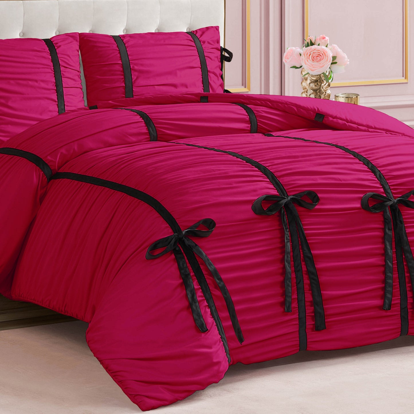 The bed features the Amelie Bows Ruched Comforter Set in bright pink with black velvet bows, matching pink pillows, and a white tufted headboard; a vase of light pink roses is on a table in the background.