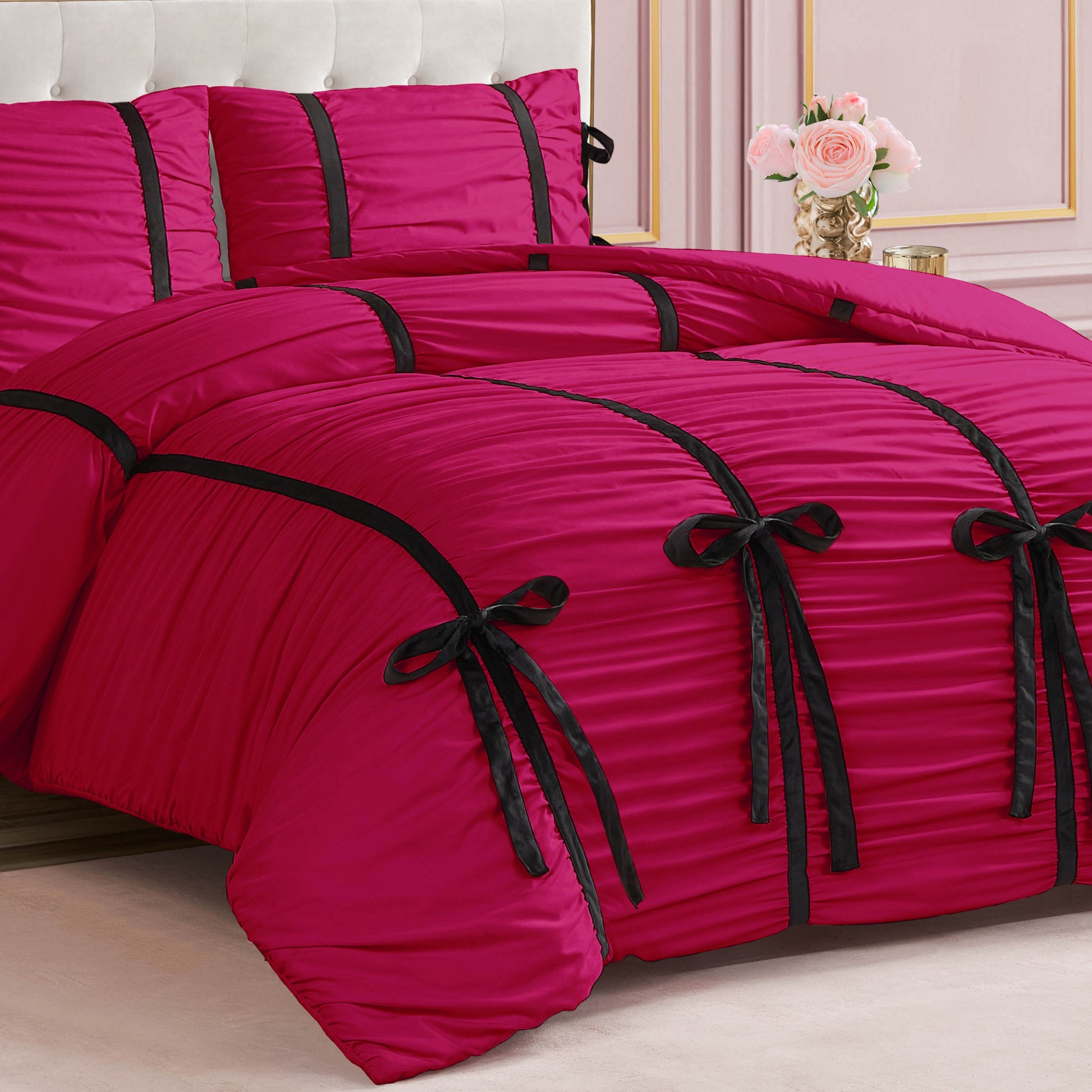 The bed features the Amelie Bows Ruched Comforter Set in bright pink with black velvet bows, matching pink pillows, and a white tufted headboard; a vase of light pink roses is on a table in the background.