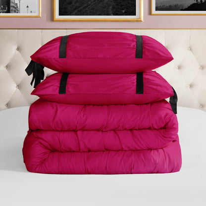 The Amelie Bows Ruched Comforter Set in bright pink features two pillows with chic black velvet bows and a folded comforter, arranged on a white bed with a tufted beige headboard and framed photos in the background.