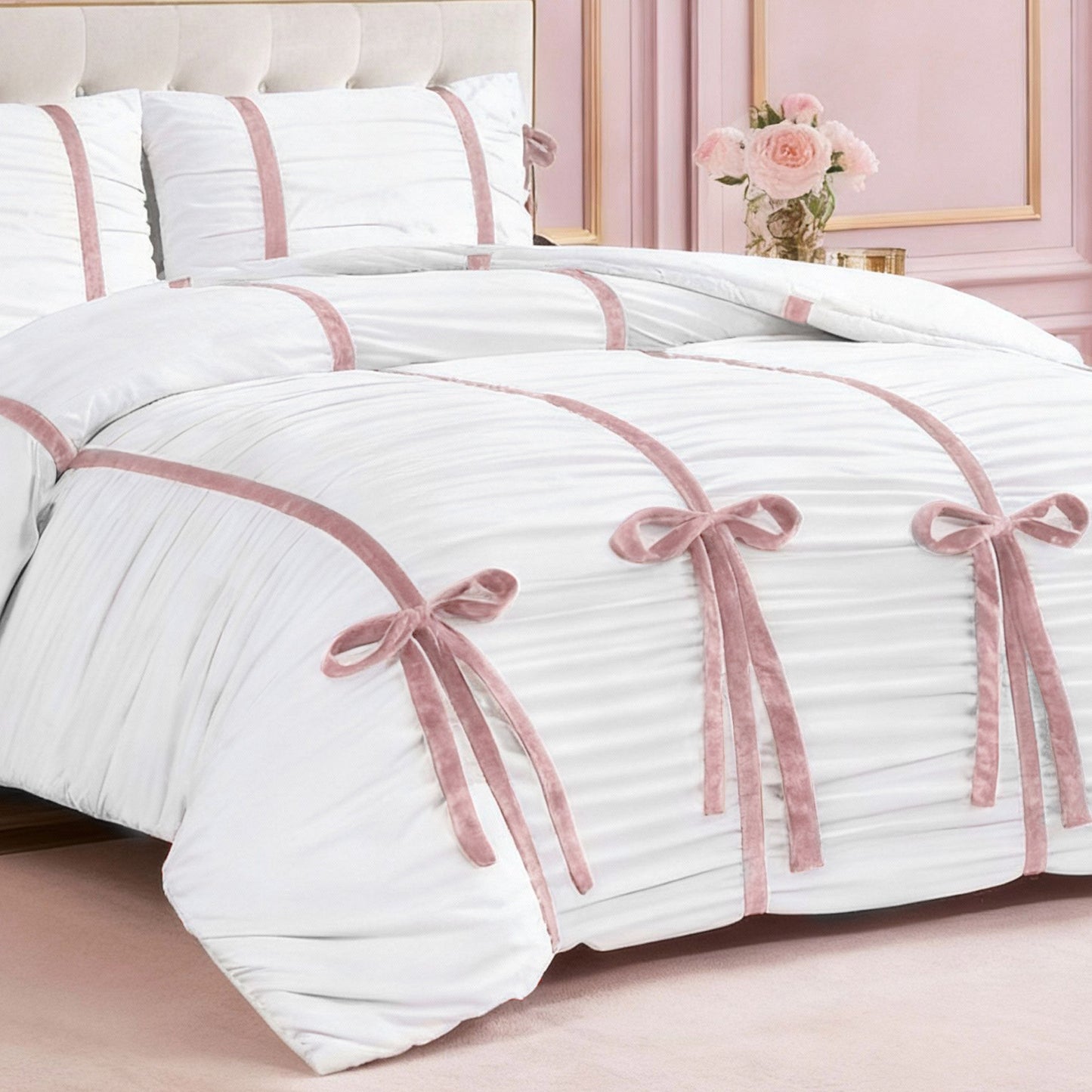 The Amelie Bows Ruched Comforter Set, featuring white fabric with pink ribbon accents and velvet bows, is neatly arranged on the bed with matching pillows. A beige headboard and a vase of flowers sit beside it against a pale pink wall.