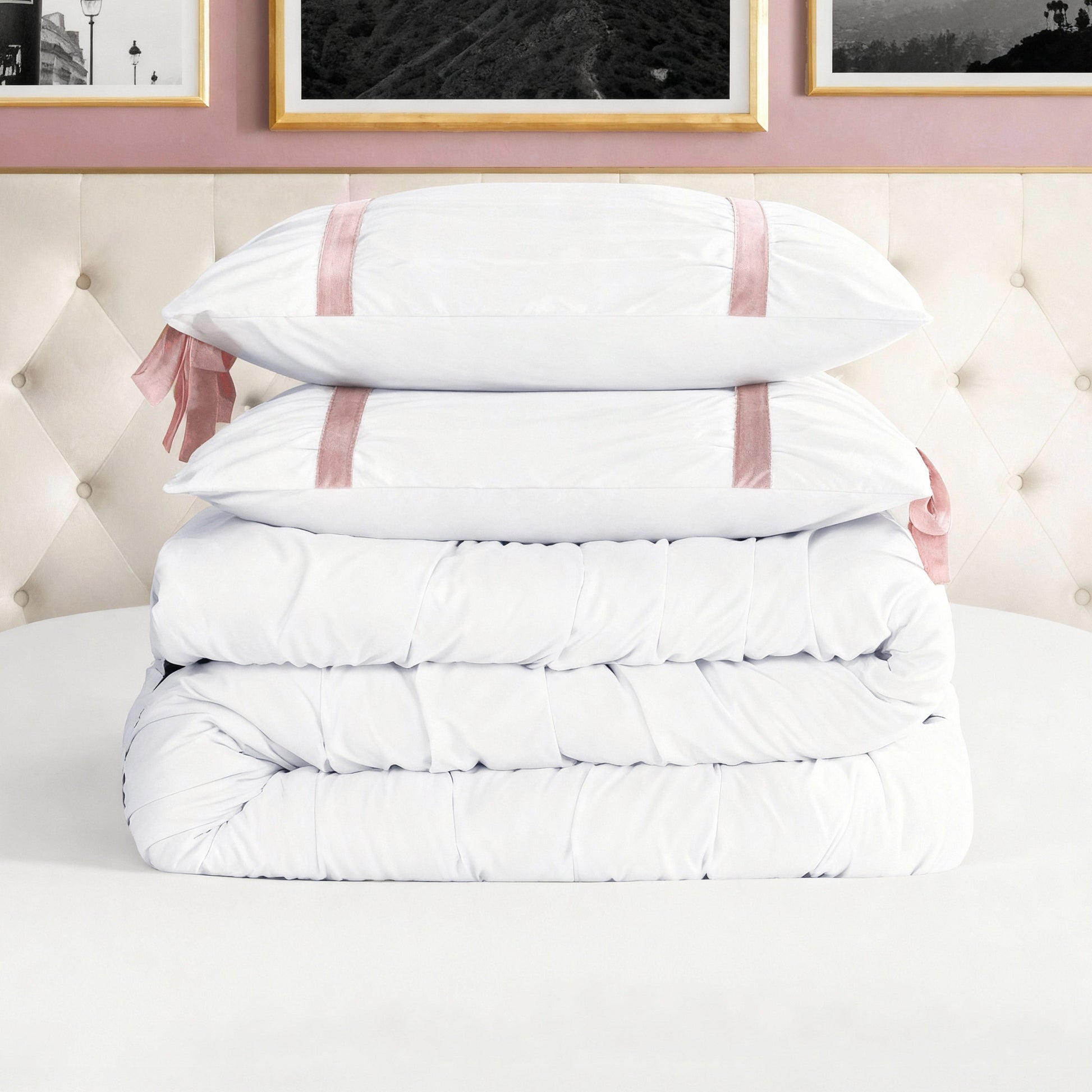 The Amelie Bows Ruched Comforter Set, including an ultra-soft folded comforter and two white pillows with pink satin ribbons and velvet bows, is displayed on a round white table in a stylish bedroom with a tufted beige headboard.