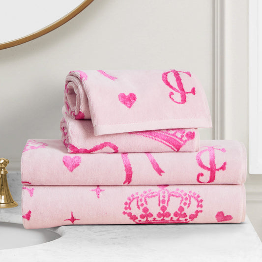 Three soft pink Printed Jacquard Bath Towel Set pieces with bright pink heart, crown, and letter motifs are neatly folded on a white bathroom countertop next to a gold faucet and a round mirror.