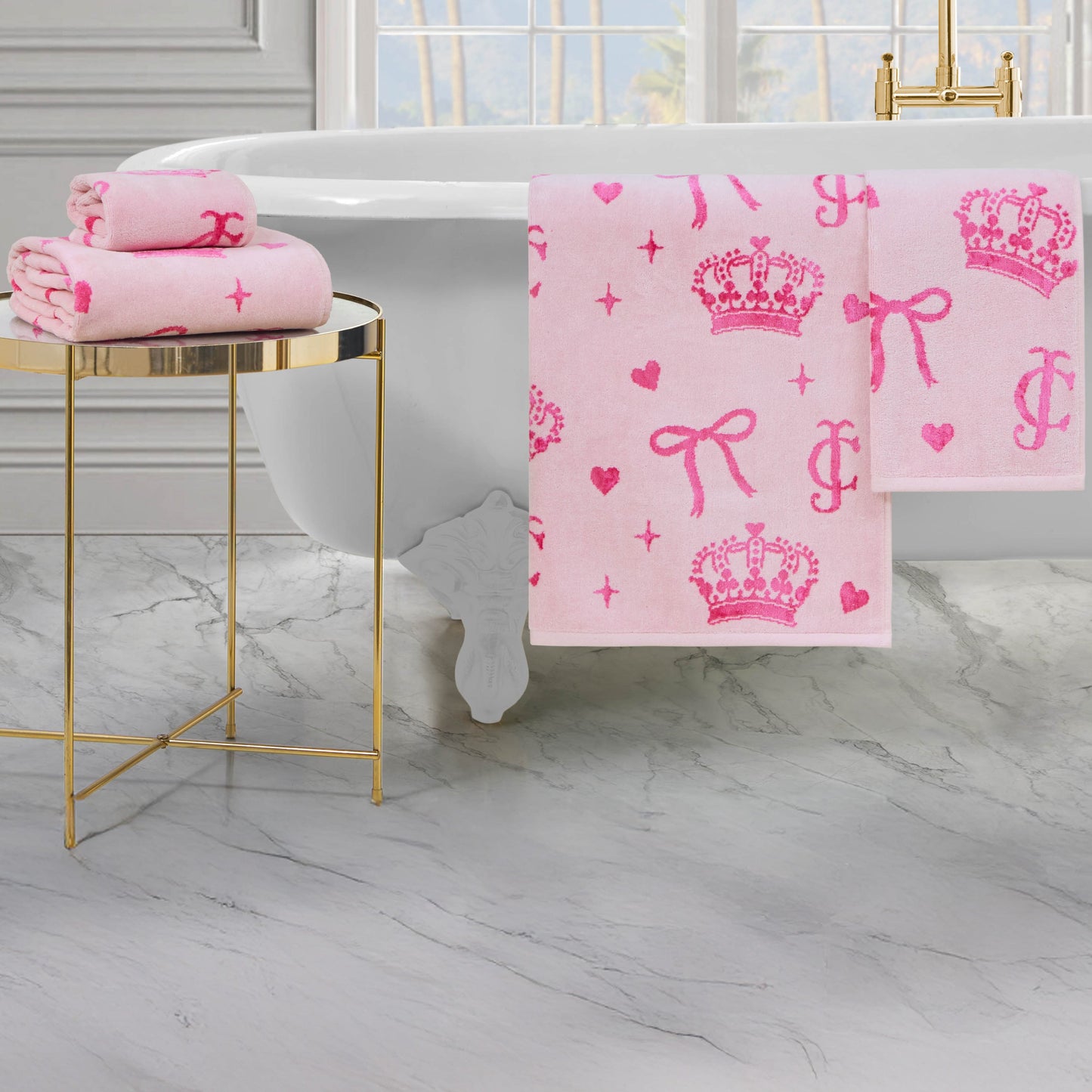 A Printed Jacquard Bath Towel Set in pastel pink with crowns, bows, hearts, and “JC” initials in bright pink sits on a gold side table and hangs over a white bathtub in a marble bathroom.