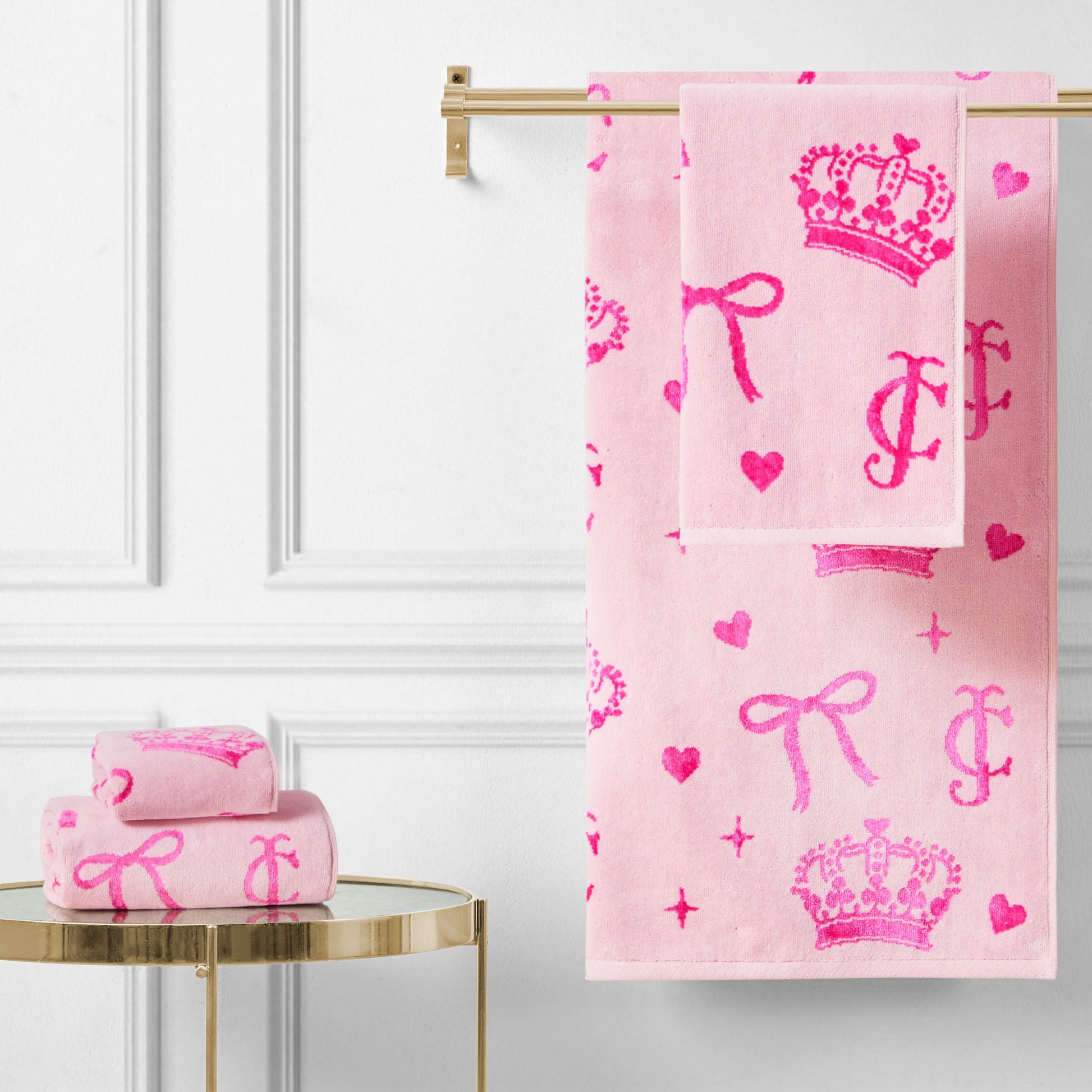 The Printed Jacquard Bath Towel Set, featuring bright pink crown, heart, bow, and letter designs, hangs on a gold rack with matching towels folded on a round gold table against a white paneled wall.