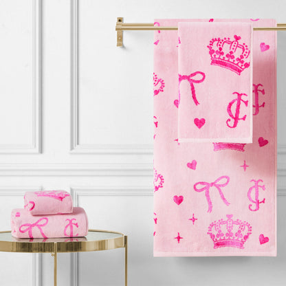 The Printed Jacquard Bath Towel Set, featuring bright pink crown, heart, bow, and letter designs, hangs on a gold rack with matching towels folded on a round gold table against a white paneled wall.