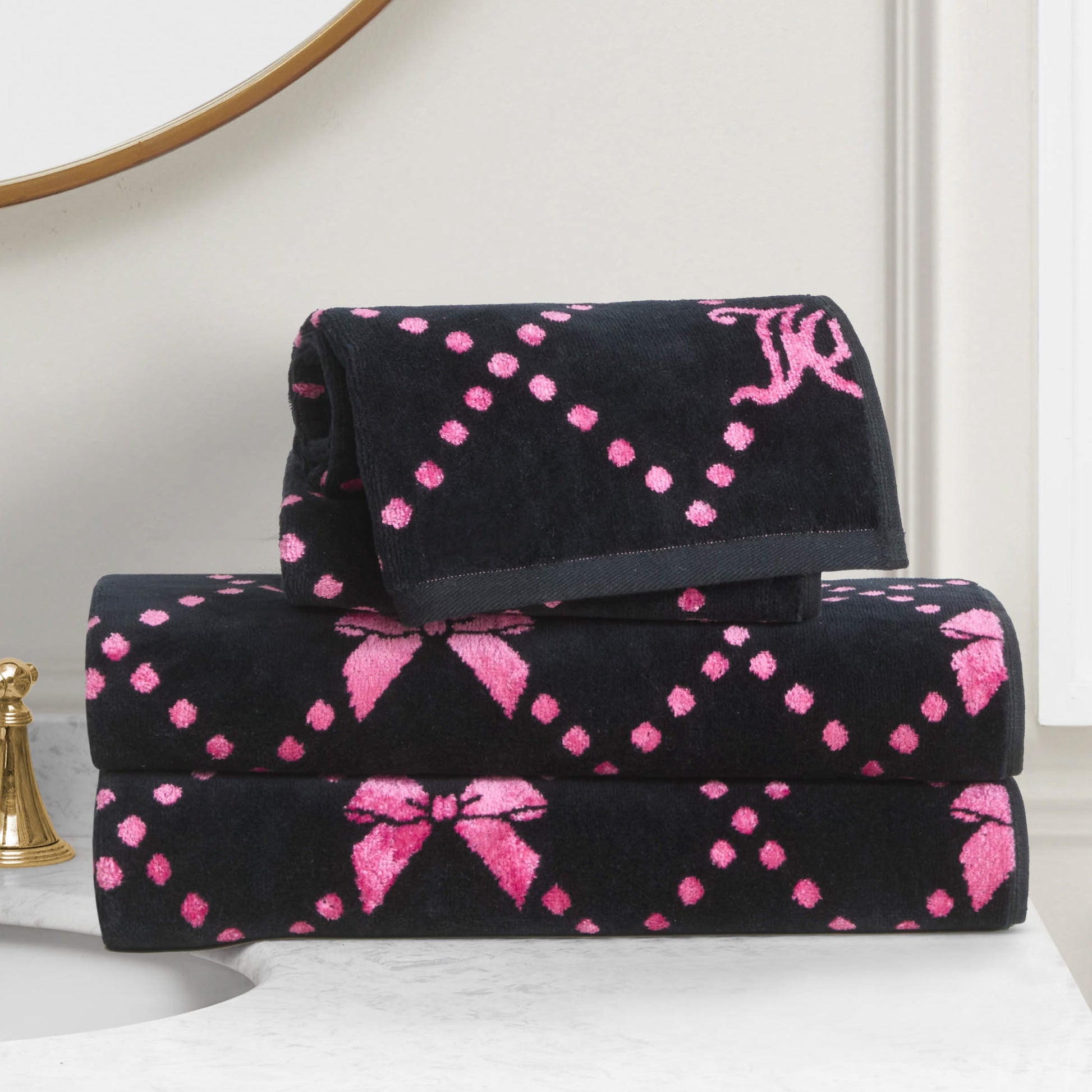 Three folded Printed Jacquard Bath Towel Set towels featuring a pink dotted diamond pattern and pink bows are stacked on a white bathroom countertop, with a gold faucet and round mirror adding to the luxurious display.