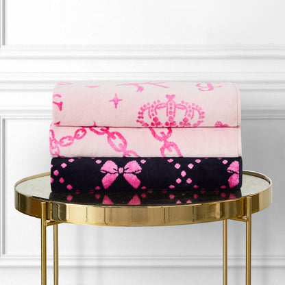 Two Printed Jacquard Bath Towels—one light pink with a crown and chain design, the other black with pink bows and diamonds—are folded on a round gold side table, set against a white panel wall.