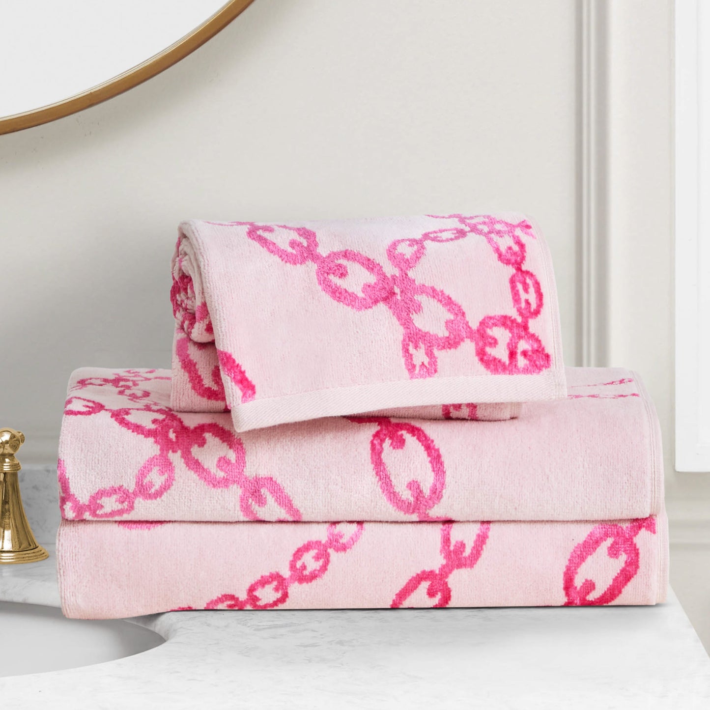 Two Printed Jacquard Bath Towel Set towels, featuring light pink with bold chain-link designs, are neatly folded and stacked on a white marble bathroom countertop beside a gold faucet.