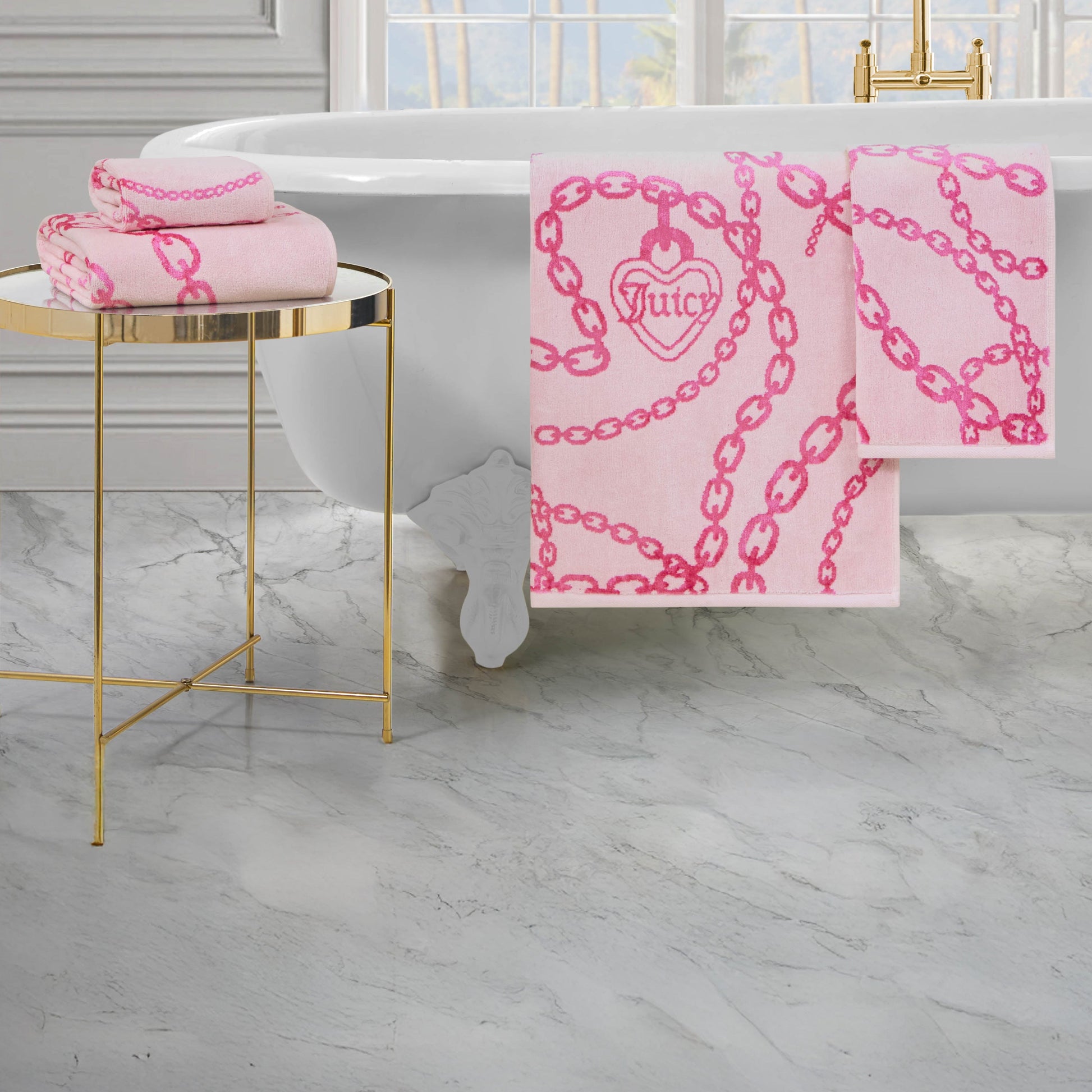 The Printed Jacquard Bath Towel Set, featuring a chain and heart design, drapes over a white claw-foot tub, with additional luxury towels folded on a gold side table beside marble flooring and gold fixtures.