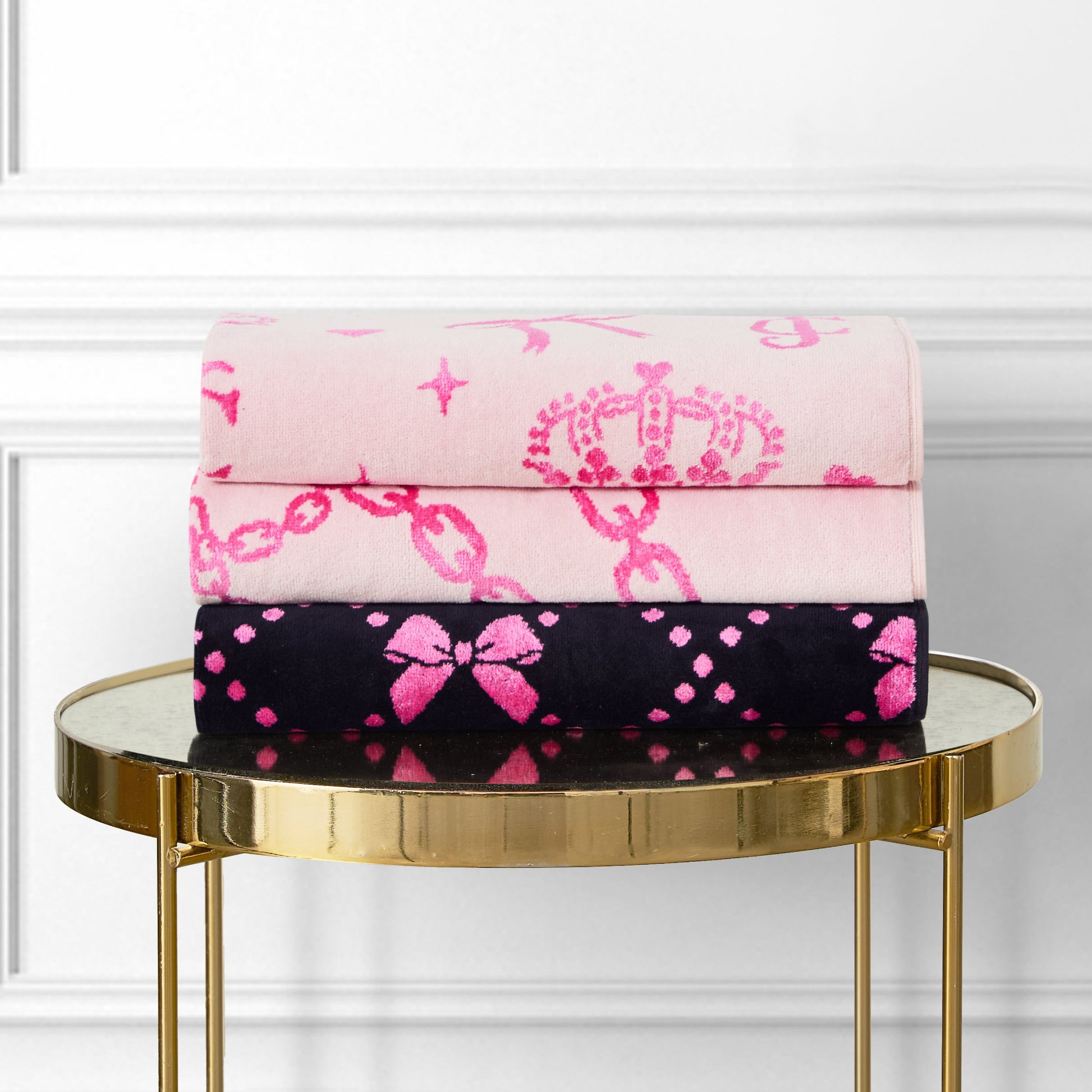 Two folded towels from the Printed Jacquard Bath Towel Set—one light pink with crowns and chains, one black with pink bows and dots—are stacked on a round gold table for a chic, luxurious bathroom display.
