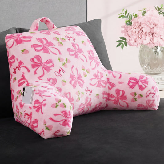 The Backrest Pillow features a handle, bows, strawberries, and “Juicy” in cursive. An iPhone is stored in its phone pocket. The pillow is on a dark sofa beside a vase of pink flowers.
