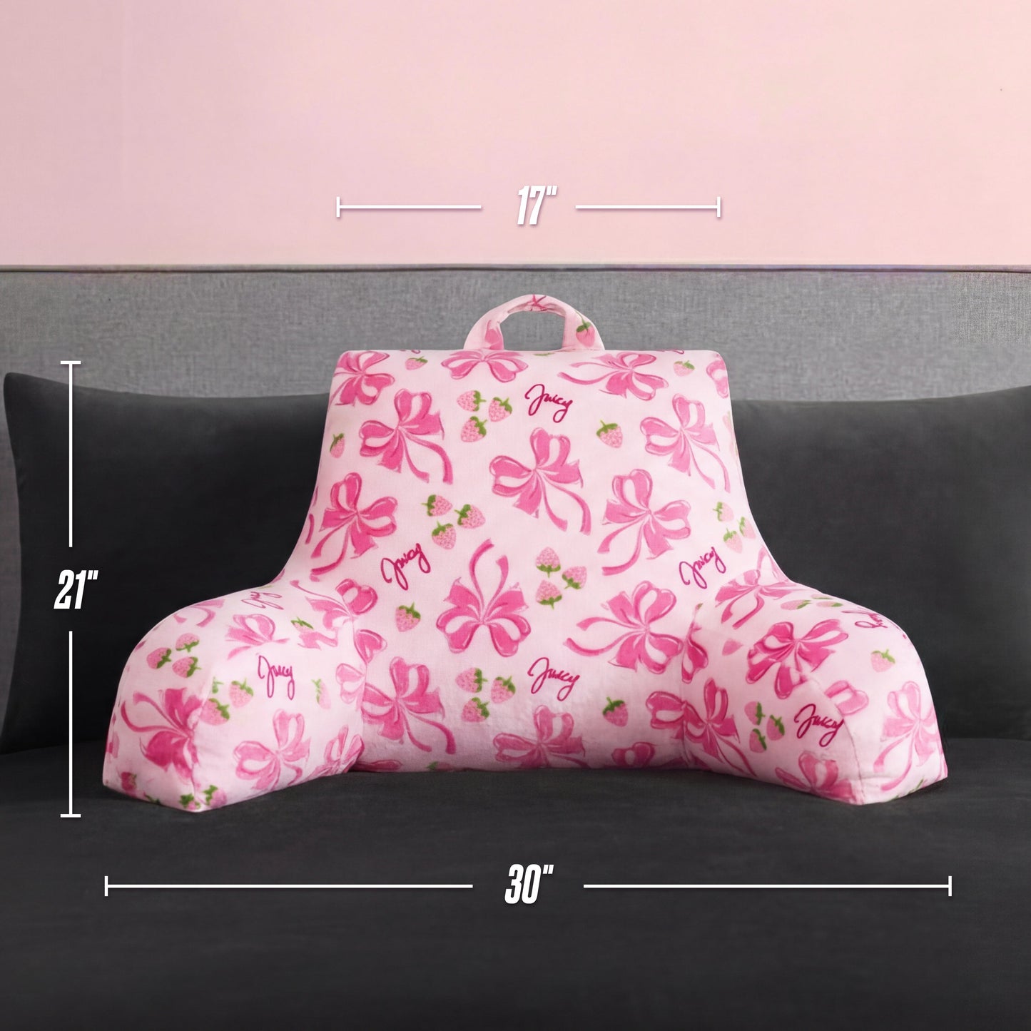 A pink Backrest Pillow with armrests, bows, strawberries, and "juicy" text rests on a gray couch. It measures 17" wide (top), 21" high, and 30" wide (bottom)—perfect for plush lounging comfort.