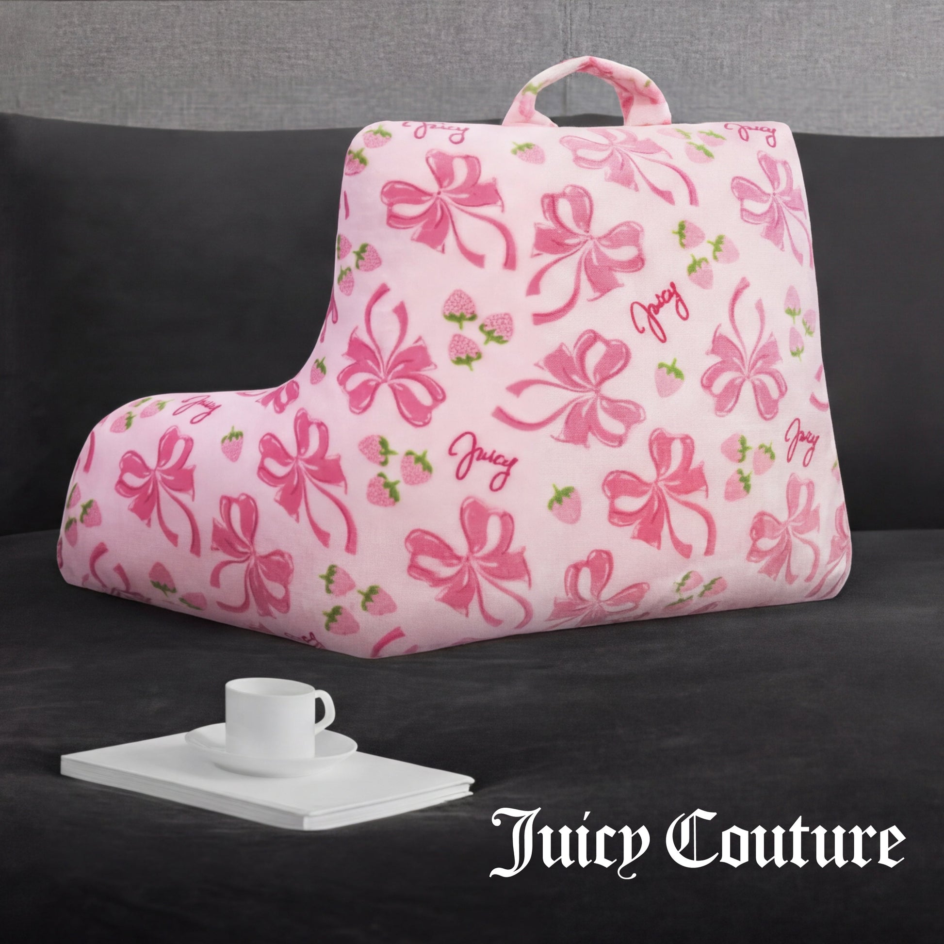 A pink Juicy Couture Backrest Pillow with bows, strawberries, and "Juicy" text rests on a dark couch beside a tray with a white teacup and saucer. The Juicy Couture logo is in the bottom right corner.