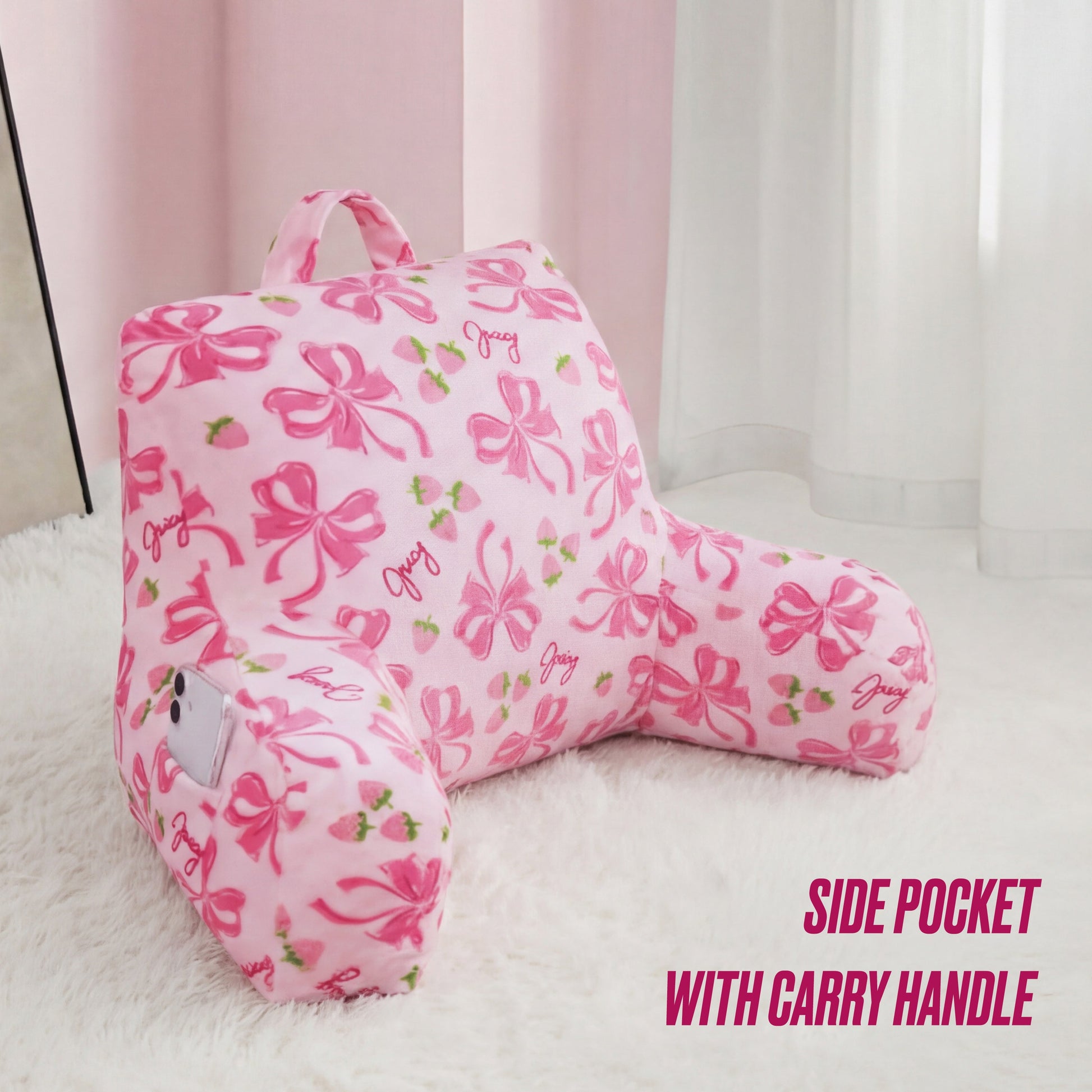 The Juicy Couture Backrest Pillow in pink features bow, strawberry, and "Juicy" designs. It has a side pocket for your phone and a carry handle. Plush and cozy, it's shown on a white furry rug near sheer curtains. Text: SIDE POCKET WITH CARRY HANDLE.