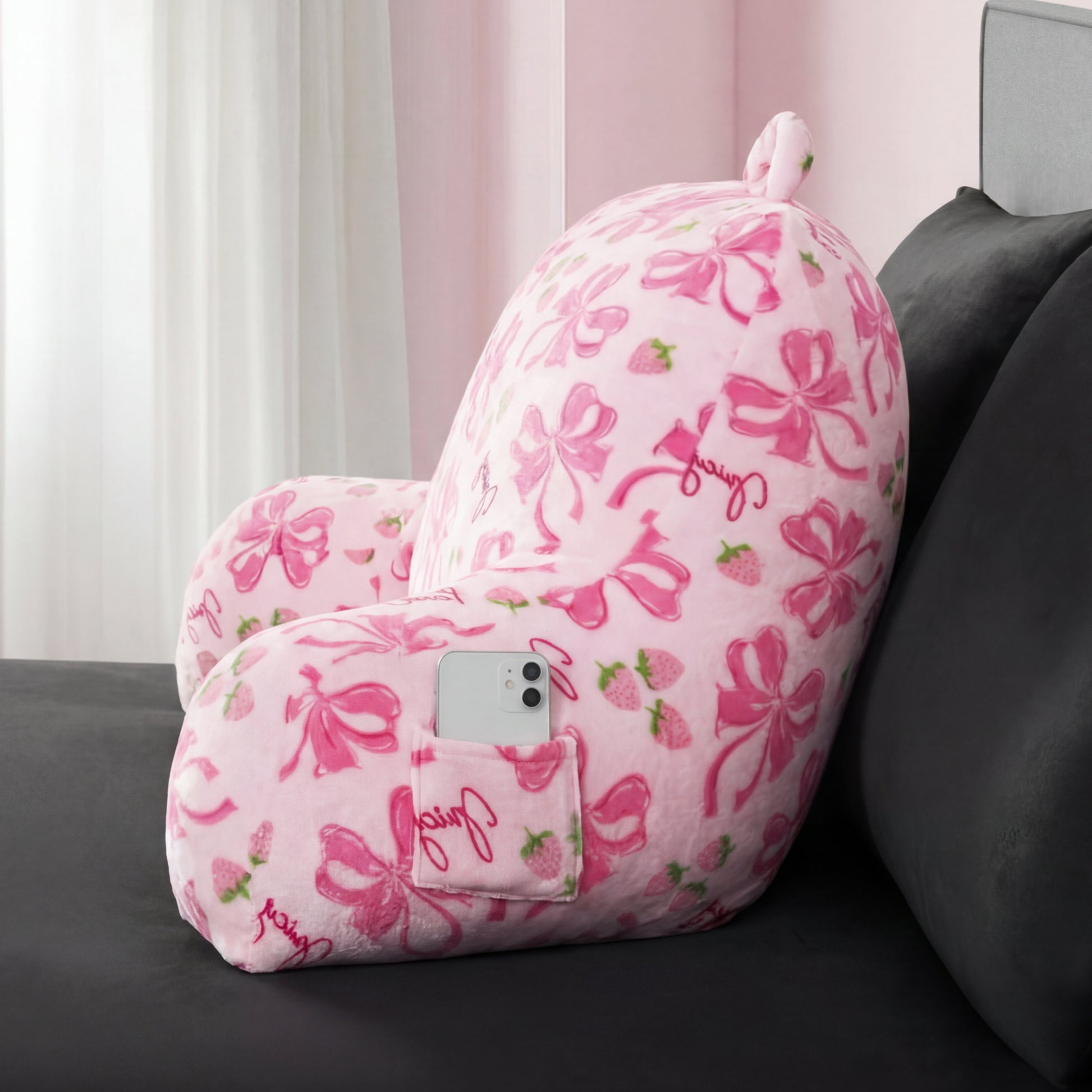 The Backrest Pillow, a plush pink pillow with phone pocket, armrests, pink bows, and strawberries, sits on a dark gray bed. A smartphone rests in its side pocket while sunlight streams through the nearby window.