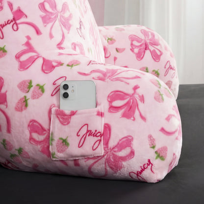 A pink Backrest Pillow with bows and strawberry print, featuring a phone pocket for your white smartphone. "Juicy" is written in cursive on the plush pillow, which sits on a dark surface with sheer curtains behind.