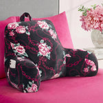 A Backrest Pillow with armrests, featuring black fabric with pink floral and chain designs, sits on a pink bed. A smartphone is stored in its side pocket, and a vase of pink flowers is placed on the nearby nightstand.