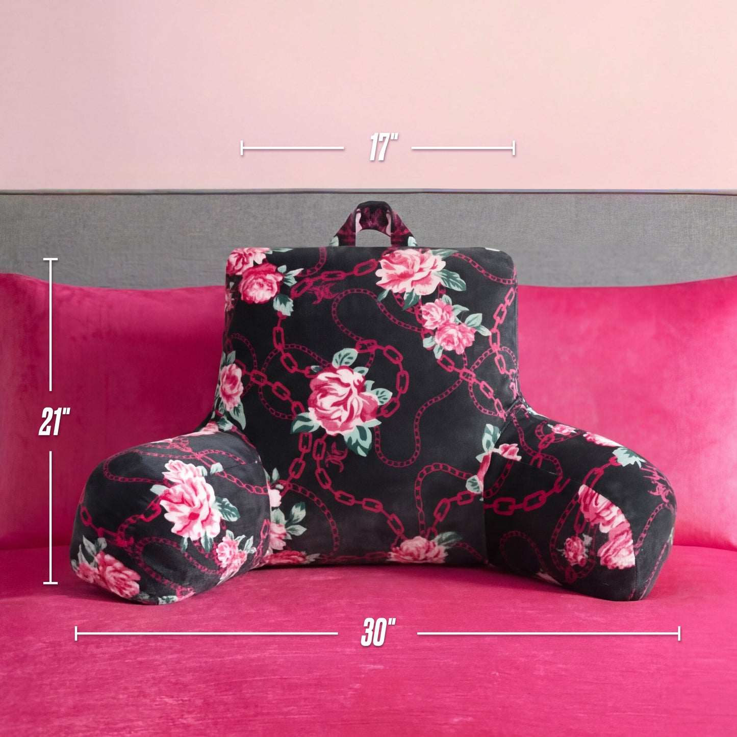 A Juicy Couture Backrest Pillow with a floral pattern and armrests rests on a hot pink bed. Measuring 17" high, 21" armrest height, and 30" wide between armrests, it is set against a light pink wall.