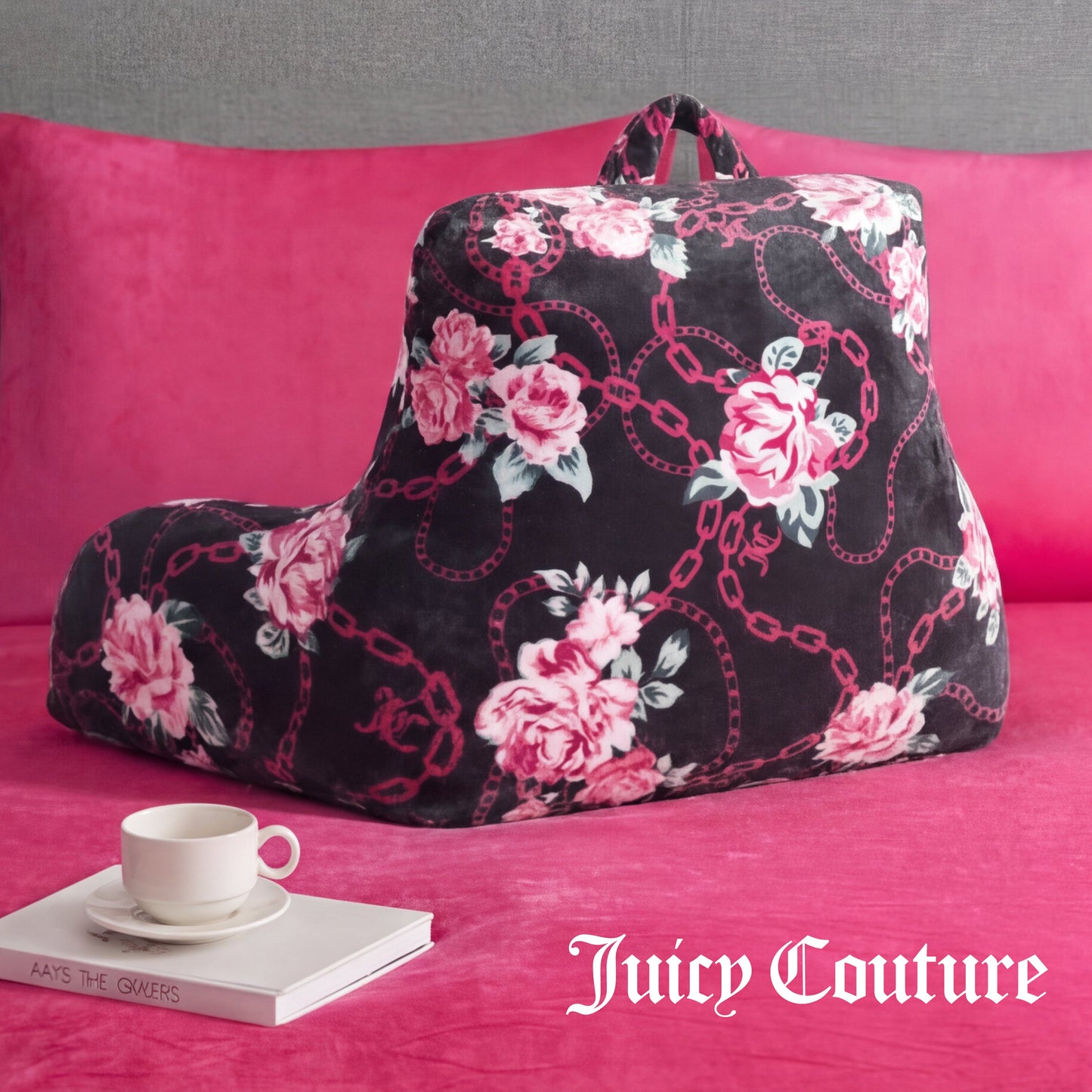 A Juicy Couture Backrest Pillow with a floral pattern rests on a pink bed, accompanied by a white teacup, saucer, and book. "Juicy Couture" is displayed in white text at the bottom right corner.