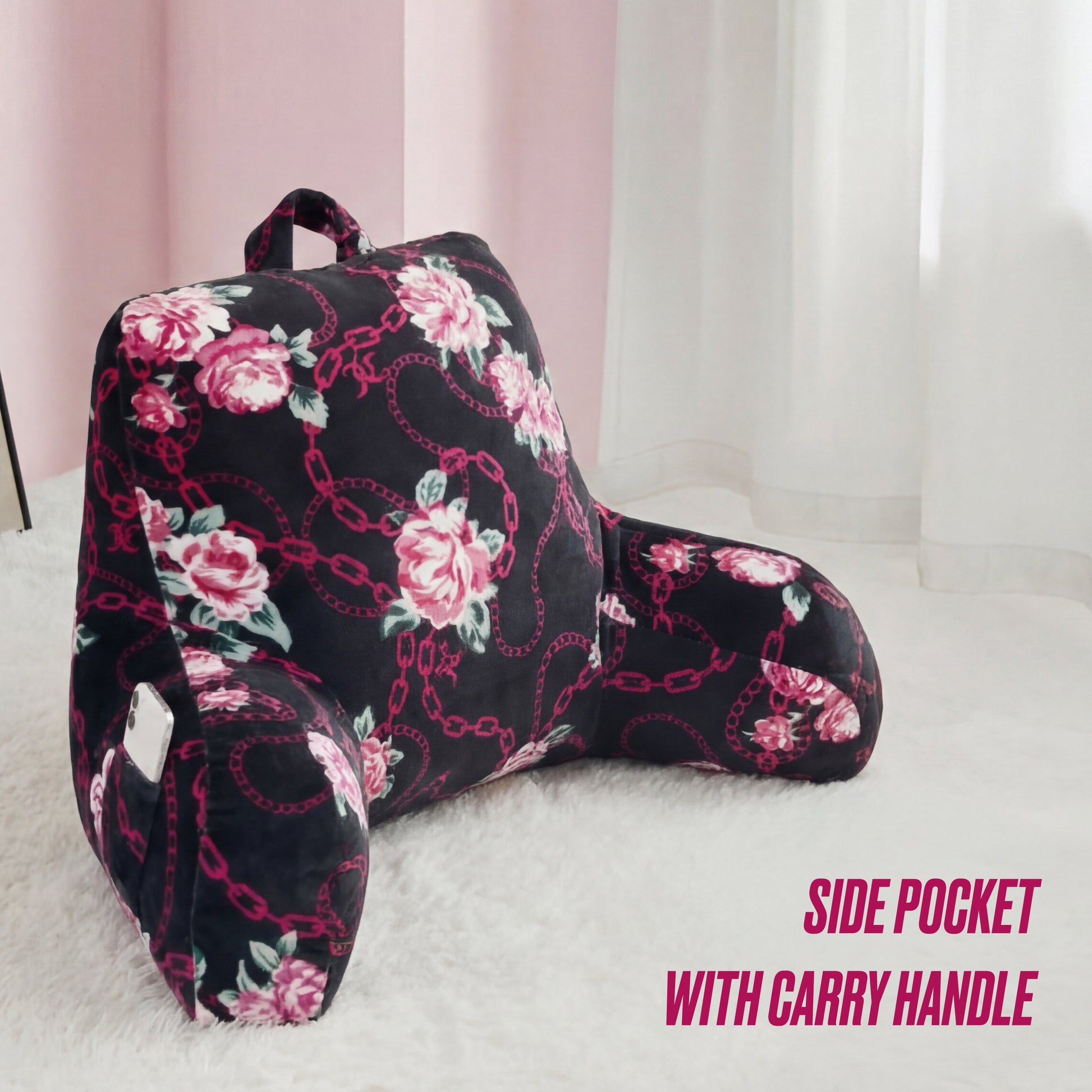 A Juicy Couture Backrest Pillow with floral design, armrests, a top carry handle, and a convenient side pocket for your phone is placed on a fluffy white rug. Text: Side pocket with carry handle.