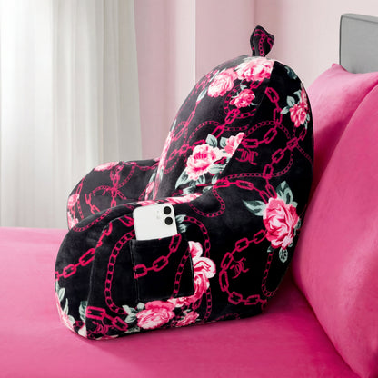 A Juicy Couture Backrest Pillow sits on a pink bed, featuring plush armrests, a pocket with a white smartphone, and a pink and black design with rose and chain patterns.