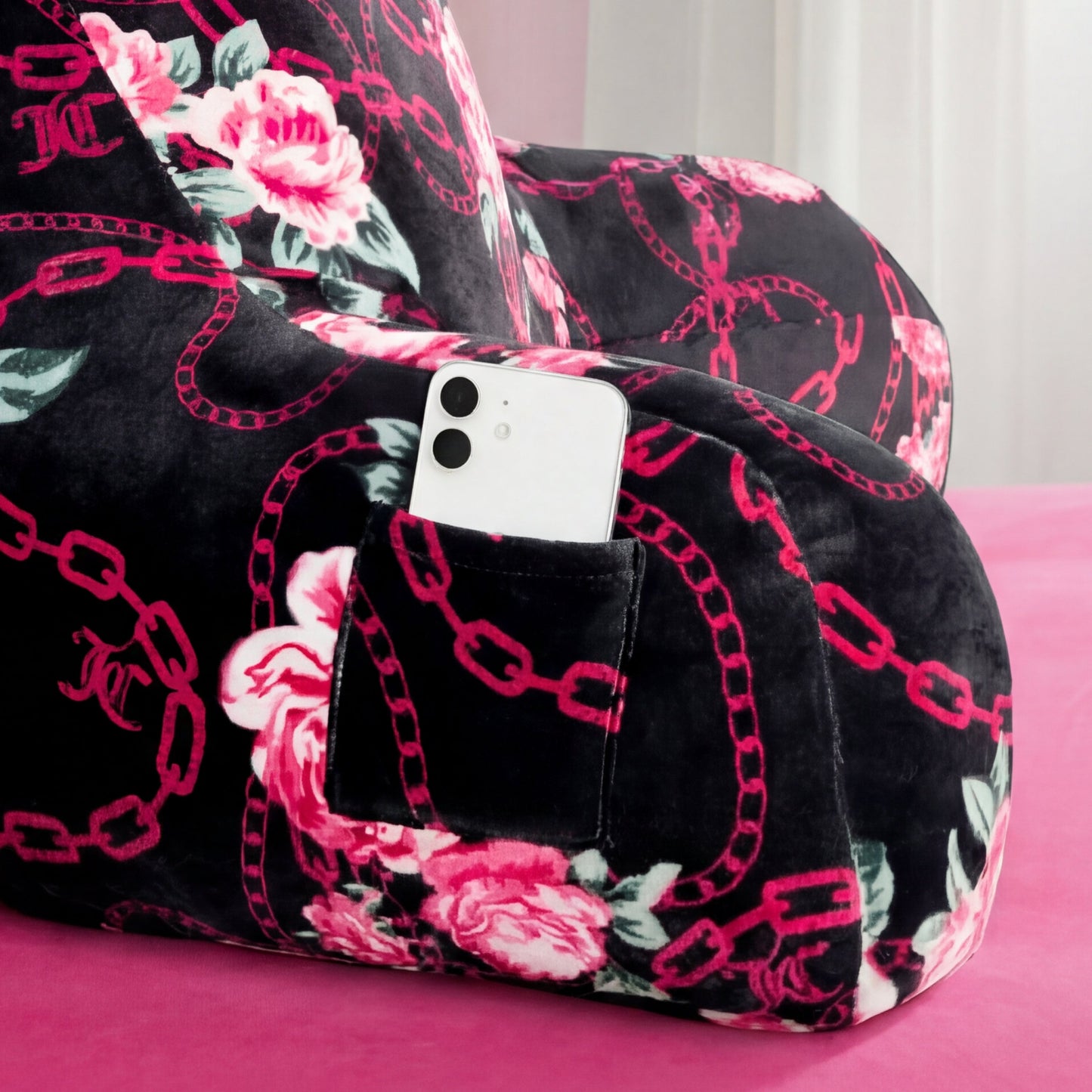 A Juicy Couture Backrest Pillow with pink chains and rose designs rests on a pink surface. Its side pocket holds a white smartphone, and white curtains are in the background.