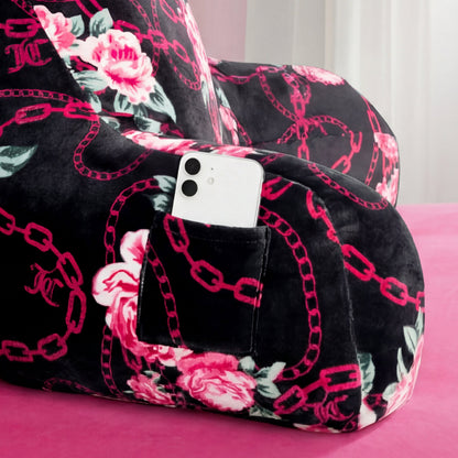 A Juicy Couture Backrest Pillow with pink chains and rose designs rests on a pink surface. Its side pocket holds a white smartphone, and white curtains are in the background.