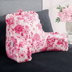 A Backrest Pillow with pink floral patterns and armrests sits on a dark grey bed, holding a smartphone in its side pocket. Pink flowers in a vase are arranged on the nightstand in the background.
