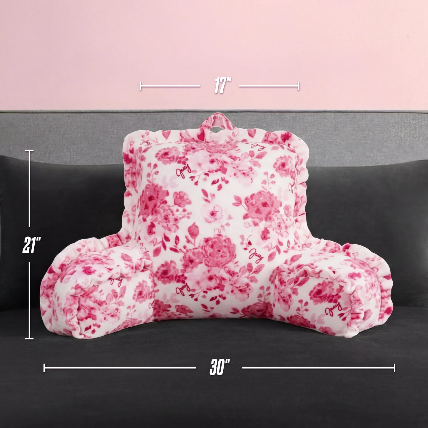 A white and pink floral-patterned Backrest Pillow with armrests is on a dark gray couch. Its overlaid dimensions are 17" tall, 21" high, and 30" wide. The background wall is light pink.
