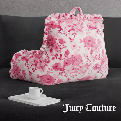 A plush Backrest Pillow with a pink floral pattern and ruffled edges sits on a dark gray couch, next to a tray holding a white cup and saucer. "Juicy Couture" appears in white text at the bottom right corner.