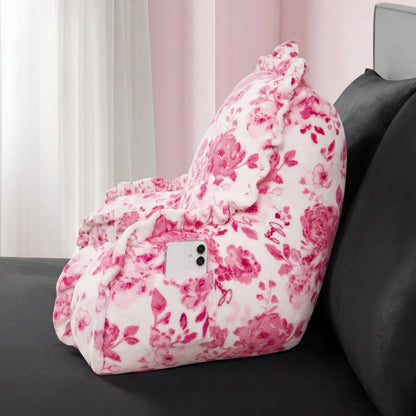 The Backrest Pillow with a floral pattern and pink ruffles rests on a dark bed, featuring a side pocket holding a smartphone, while sheer curtains brighten the background.