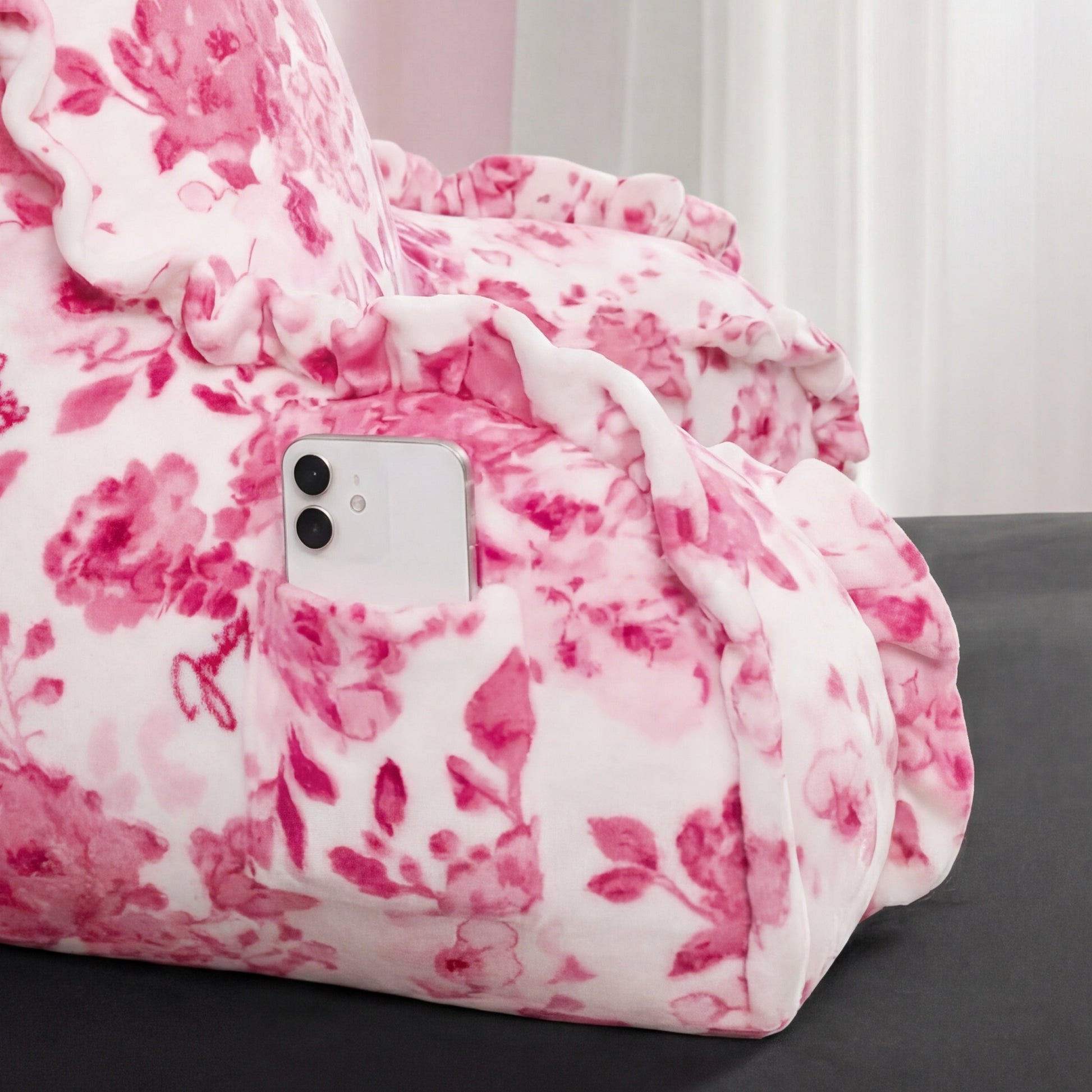 A white smartphone sits in the side pocket of a Backrest Pillow with pink floral patterns and ruffled edges, set against a soft, cozy background.