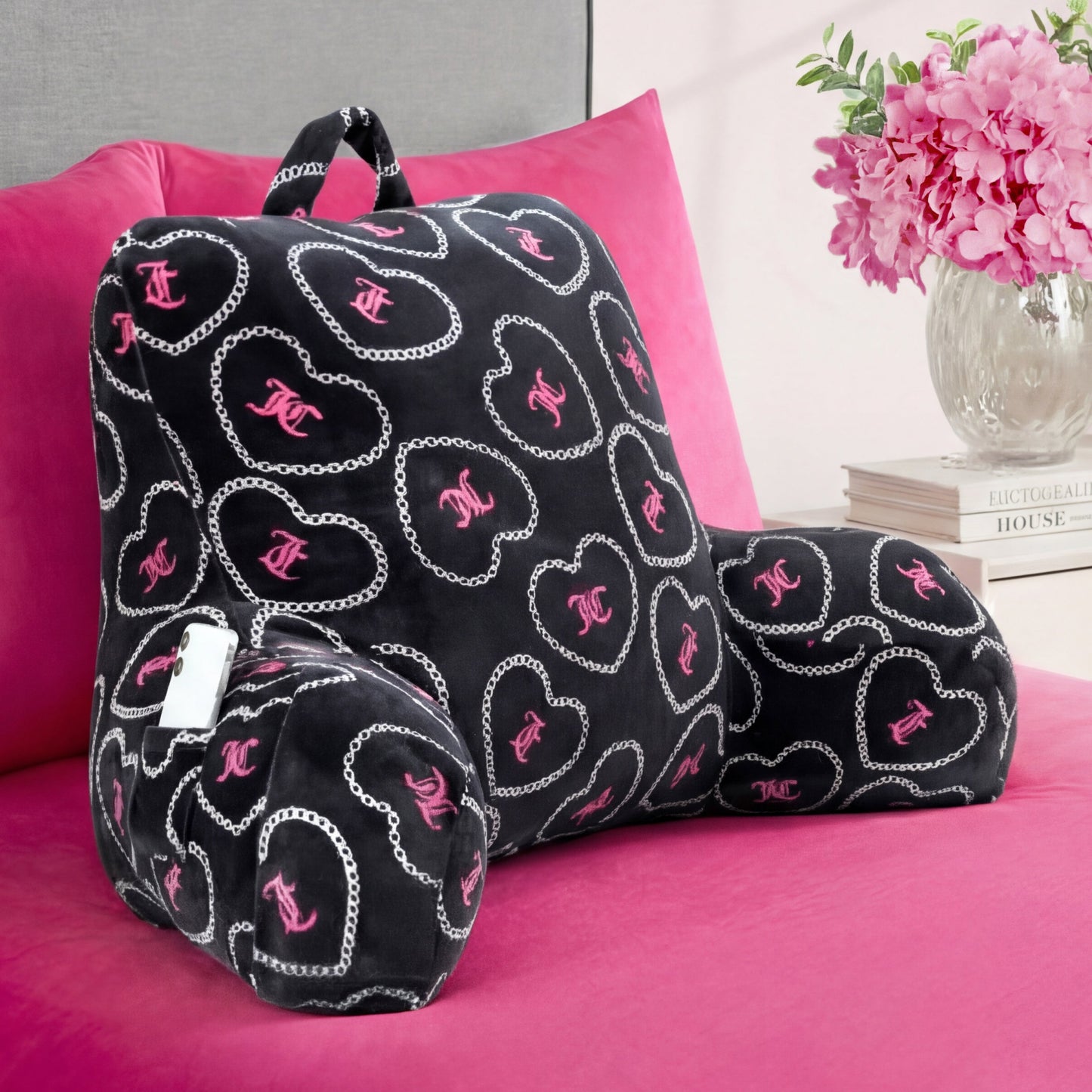 A Backrest Pillow with pink and white heart and chain designs is placed on a hot pink bedspread, while pink flowers and books are neatly arranged on the nightstand nearby.