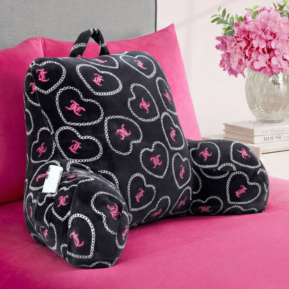 A Backrest Pillow with pink and white heart and chain designs is placed on a hot pink bedspread, while pink flowers and books are neatly arranged on the nightstand nearby.