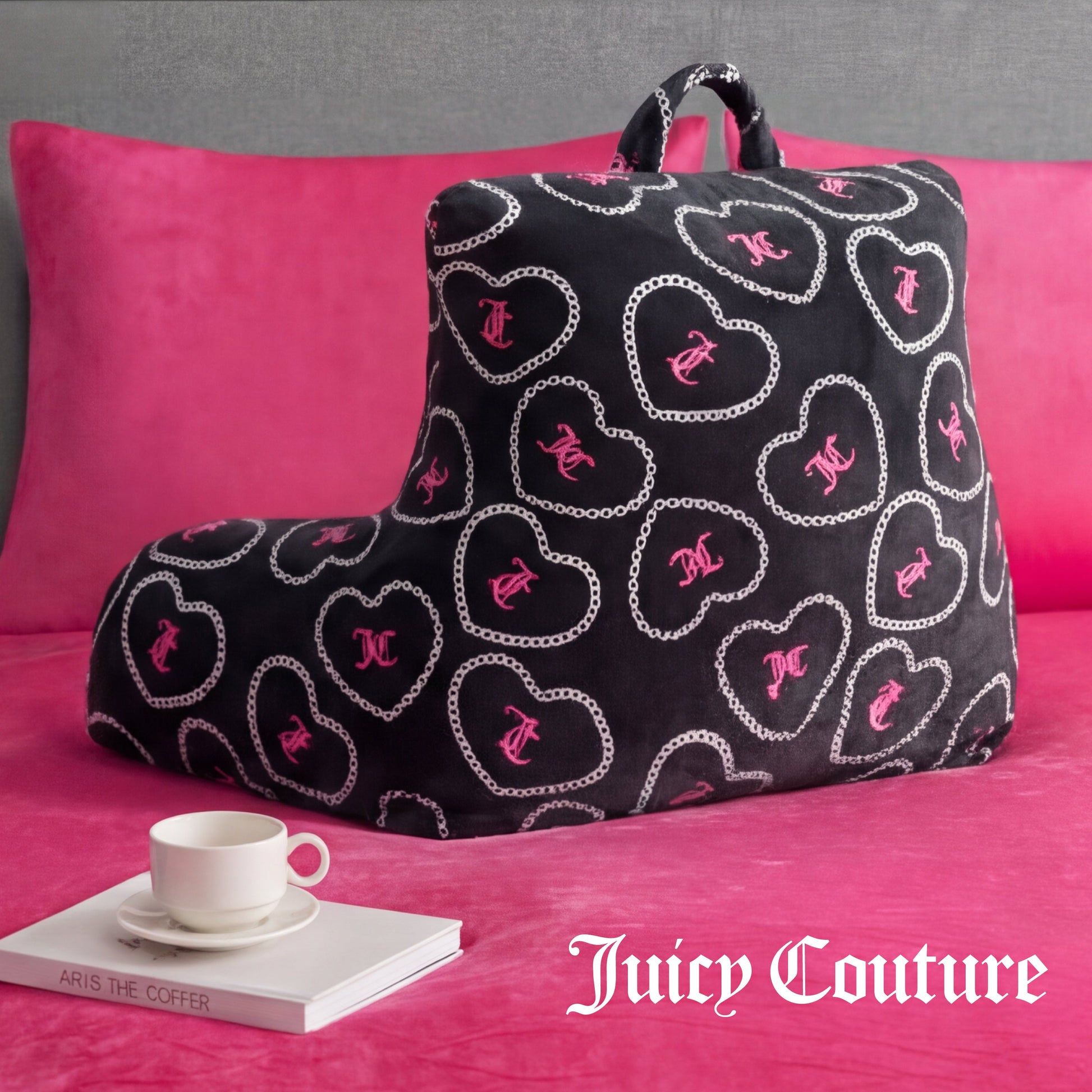 A black Backrest Pillow with pink and white heart patterns sits on a pink bed, accompanied by a white teacup and books. The Juicy Couture logo appears in the bottom right corner.