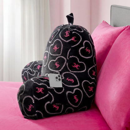 A black Backrest Pillow with silver chains and pink hearts sits on a pink bed. A smartphone is tucked in one of the pillow’s arm pockets. Sheer curtains and a gray headboard appear in the background.