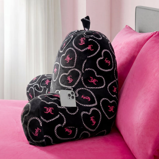 A black Backrest Pillow with silver chains and pink hearts sits on a pink bed. A smartphone is tucked in one of the pillow’s arm pockets. Sheer curtains and a gray headboard appear in the background.