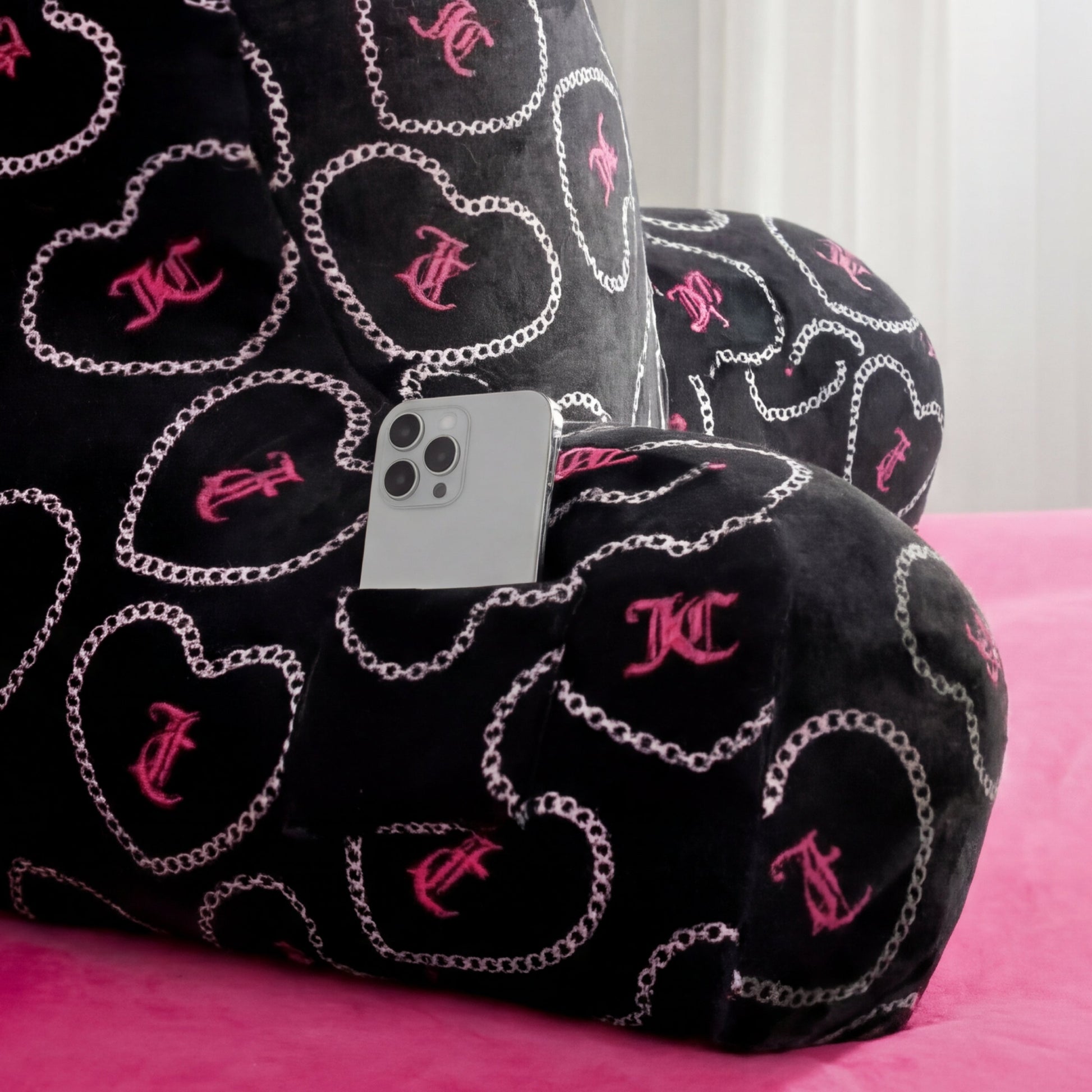The Backrest Pillow features pink heart and chain patterns, a small pocket holding a silver smartphone, and sits on a pink bedspread with a white curtain in the background.