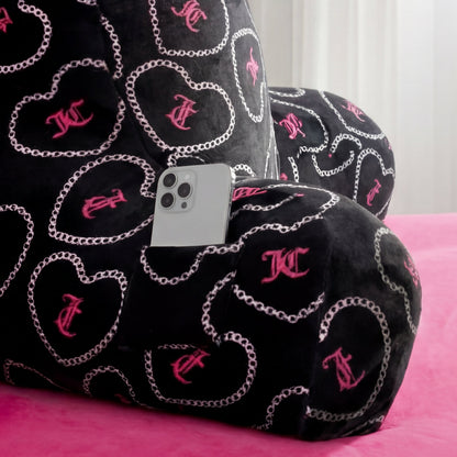 The Backrest Pillow features pink heart and chain patterns, a small pocket holding a silver smartphone, and sits on a pink bedspread with a white curtain in the background.