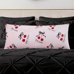 A Plush Body Pillow in light pink with red cherry and black bow designs and cursive text adds a chic pattern accent on a black tufted bedspread, set against a light-colored headboard.