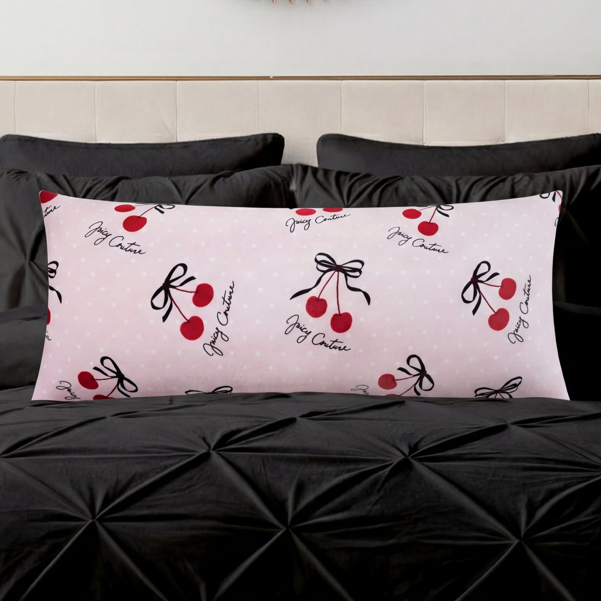A Plush Body Pillow in light pink with red cherry and black bow designs and cursive text adds a chic pattern accent on a black tufted bedspread, set against a light-colored headboard.